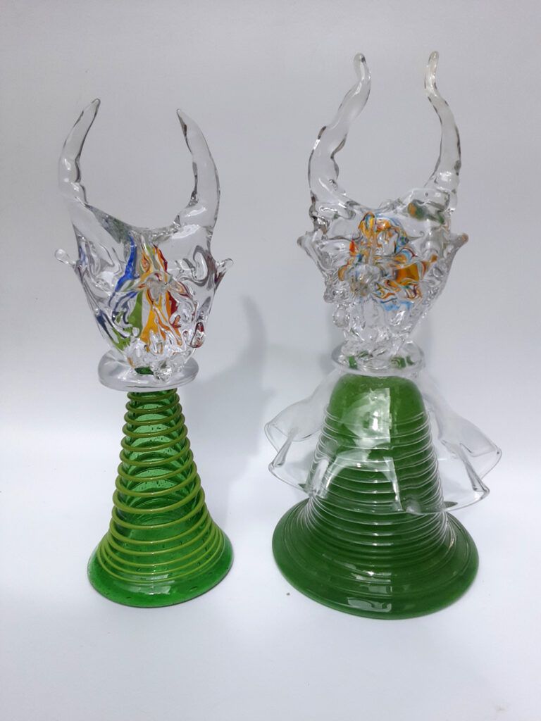 zwei Glasfiguren, bestehend aus einem grünen Fuß mit Teufelskopf aus Klarglas